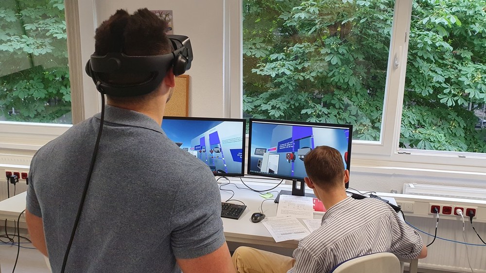 VR-Lab_Gebaeudesystemtechnik Zwei Studierende sind von hinten zu sehen, einer steht und hat eine VR-Brille auf, der andere sitzt an einem Tisch, vor ihm zwei Bildschirme, die eine VR-Umgebung zeigen..