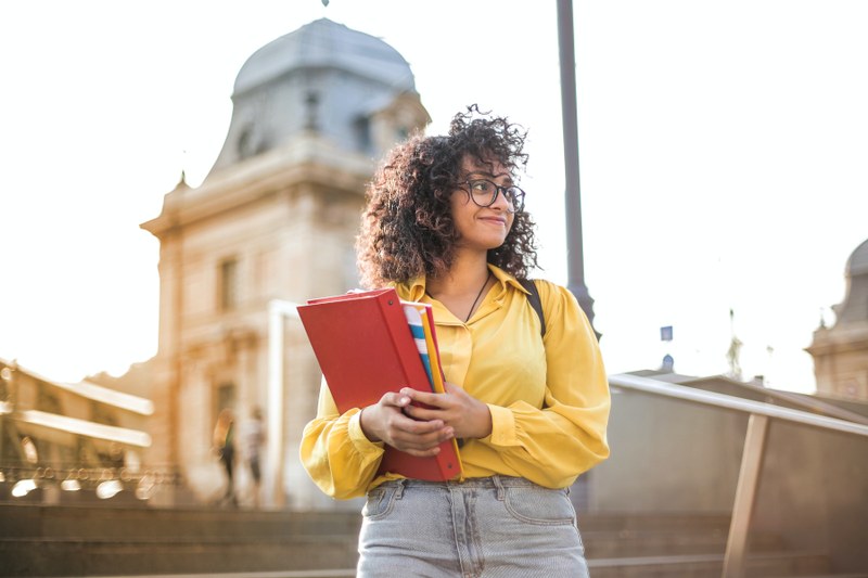 Studentin Studentin mit Ordner und Blog im Arm steht vor einem Uni-Gebäude.