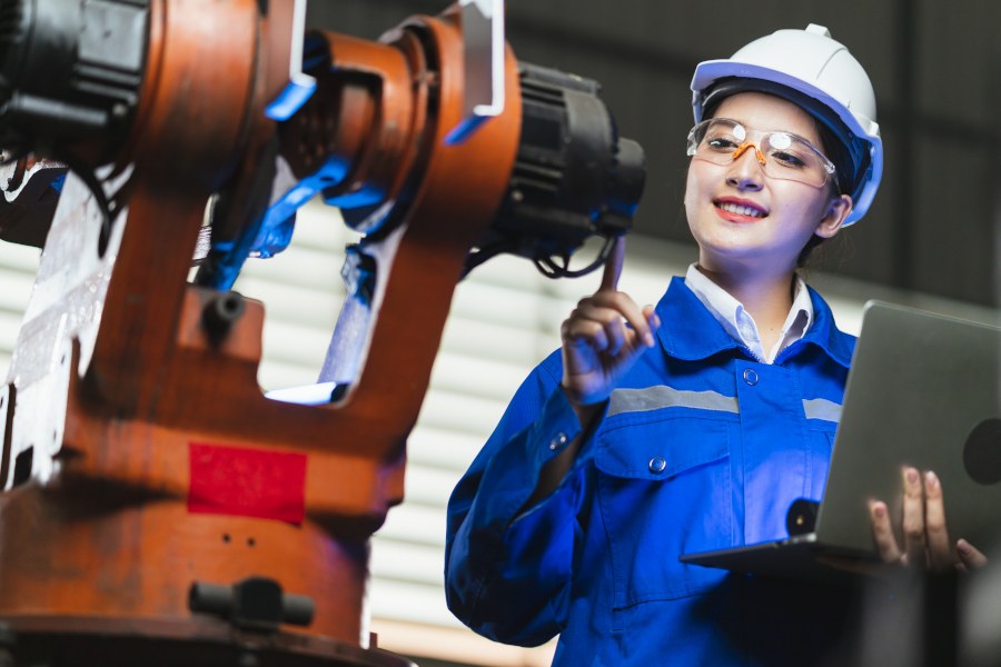 Praktikum Junge Frau in Arbeitskleidung mit Helm, Brille und Overall, hält Laptop in der Hand und inspiziert eine Maschine.